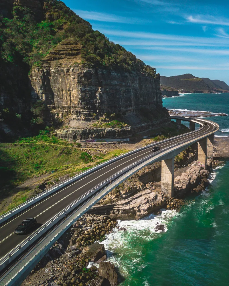 Narrow Curvy Road With Cars Alongside Green Mountains